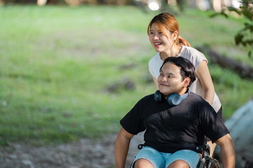 A young woman assists man in a wheelchair outdoors.